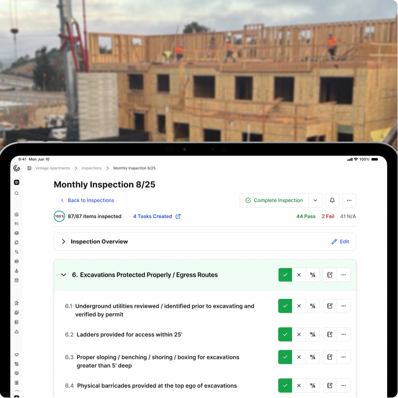 A photograph showing Constructable's Inspections feature being used at a construction site. In the foreground, we see a tablet that has Constructable open to an inspection and some items have been marked as passed as the inspection is still being performed. There are buttons for each inspection item to mark them as pass, fail, or not applicable. There are also buttons to quickly add notes when needed, or even create action items. As the inspection is performed, the tablet is showing a real-time list of the inspection items that have been completed, along with the pass rate of the inspection overall.